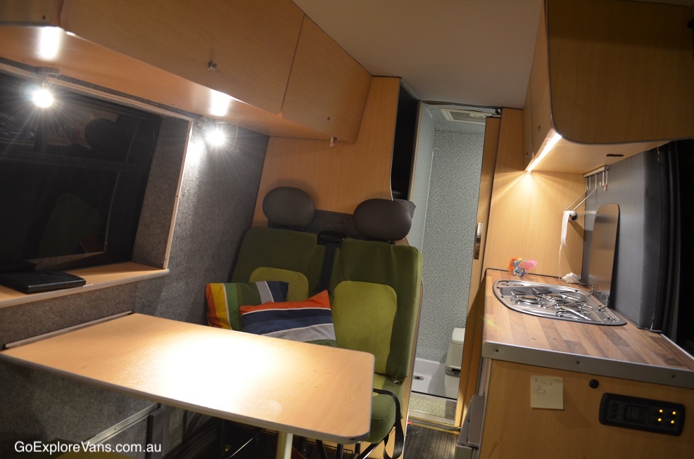 Inside a campervan showing the seating area and kitchen, with the bathroom in the rear.
