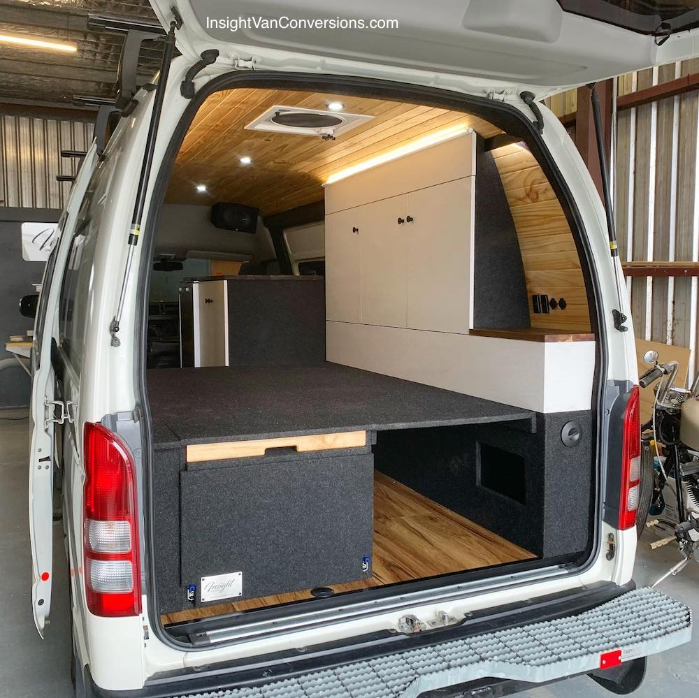 Looking into a converted campervan through the rear door, showing the bed platform and storage on the side wall of the van.