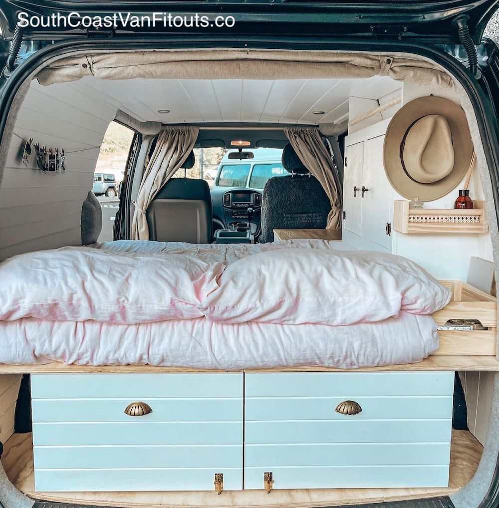 Rear door of a campervan is open showing a sleeping platform with pink fluffy duvet and storage drawers below.