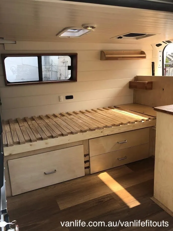 Wooden interior of a campervan showing the bed platform with storage drawers below.