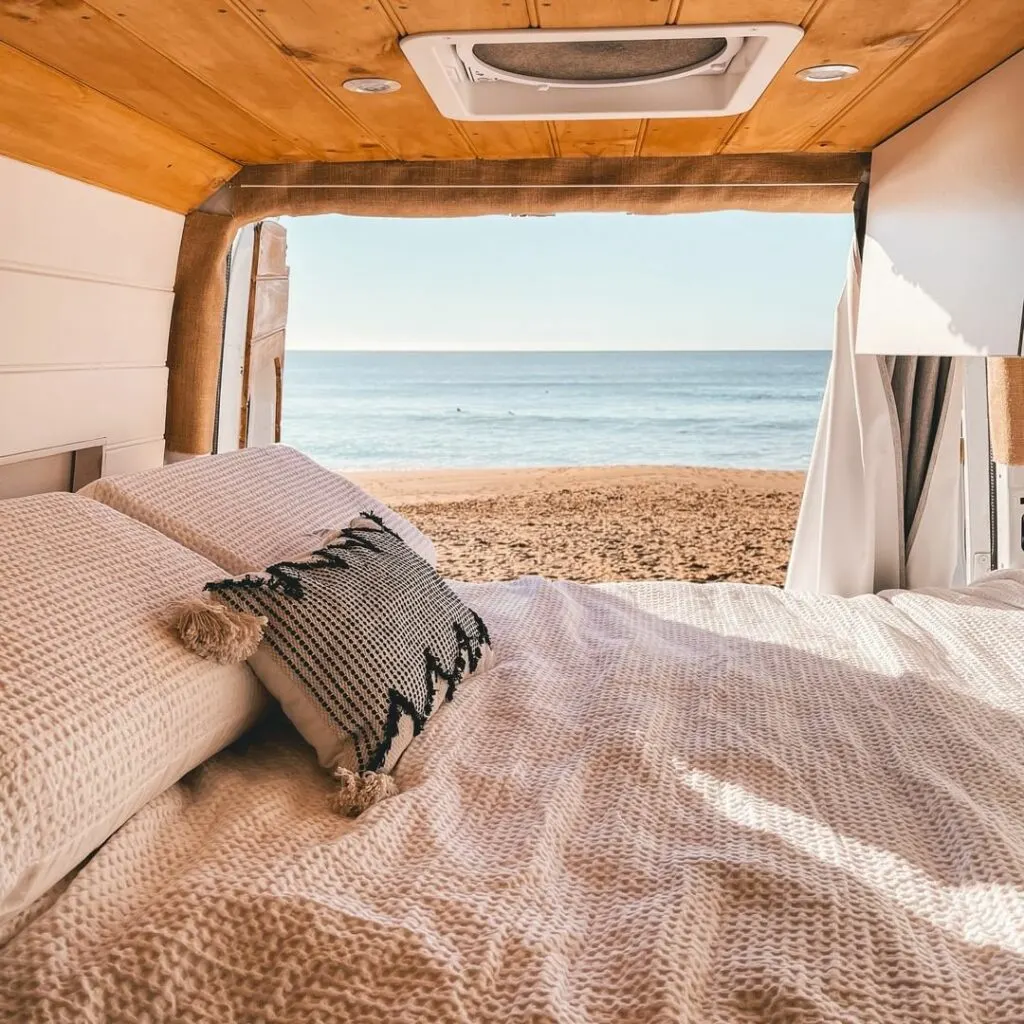 Bed inside a camper van conversion looking out the back doors to a beach.