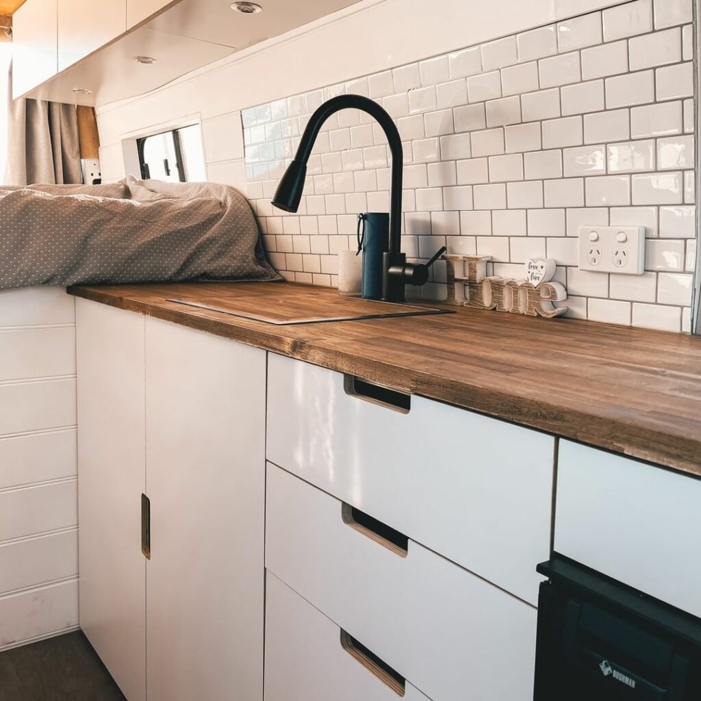 Wooden kitchen countertop with white cabinets and splashback, inside a converted campervan.