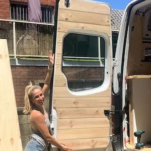 Woman standing at the back doors of a converted camper van showing off her handiwork.