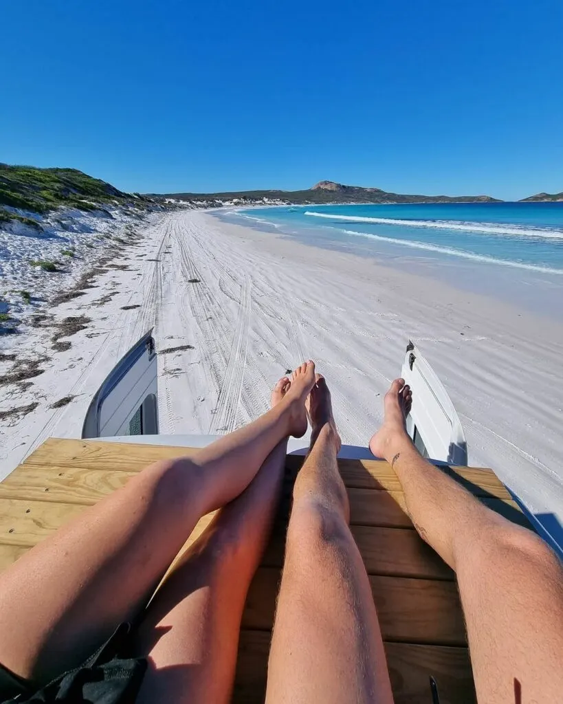 Couple sitting on the roof deck of a campervan with a long sandy beach in the background.