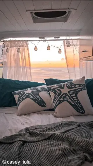 Cozy decor on the bed inside a campervan, with the back doors open and overlooking a view of the ocean and setting sun.