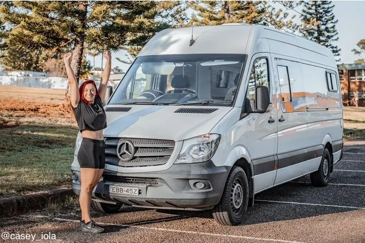 Smiling woman with her arms raised next to a white camper van.