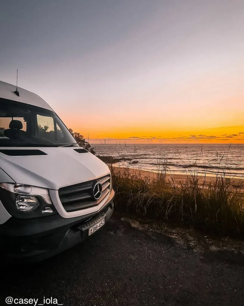 White camper van next to a beach with sun setting in the background.