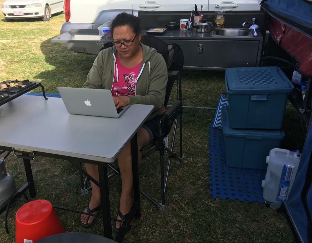 Woman working on a laptop sitting on a camp chair with camping gear around her.