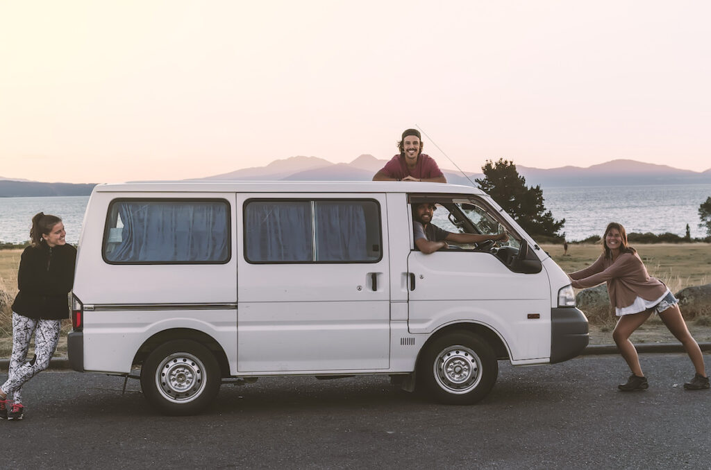 A group of 4 happy friends (2 girls and 2 boys) who love traveling stay close to their van at sunset. They travel with this simple and cheap white camper van as backpackers on a budget.