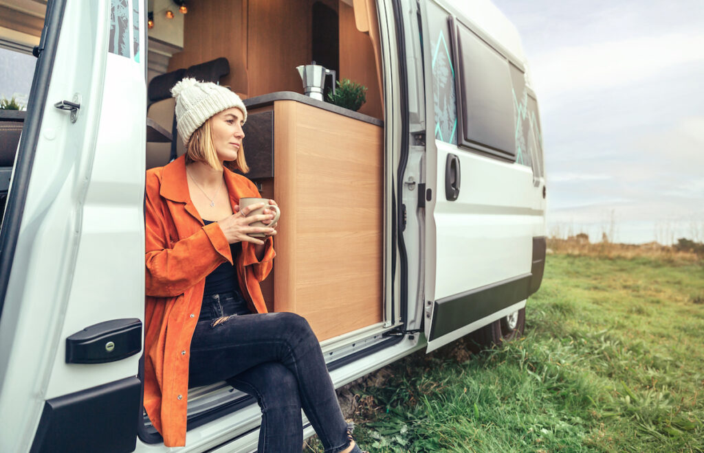 Woman drinking a coffee while sitting at the side door of a white campervan.